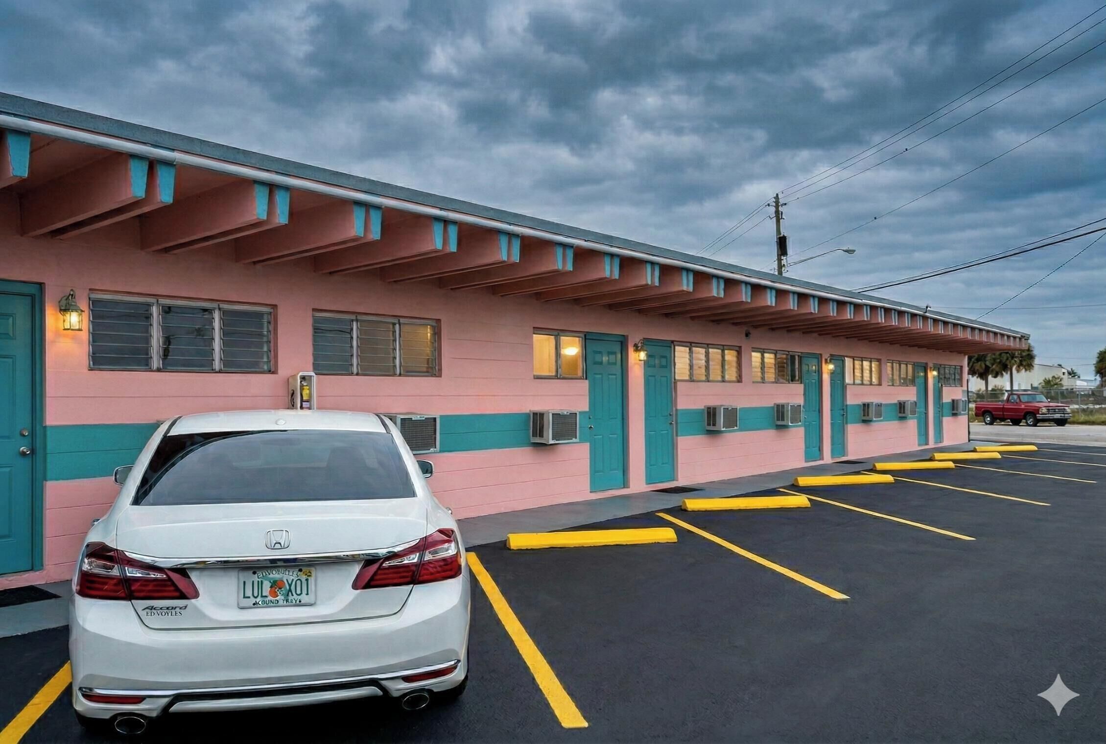 A motel with a pink and teal facade, a white car parked in the lot, and a red truck in the background.