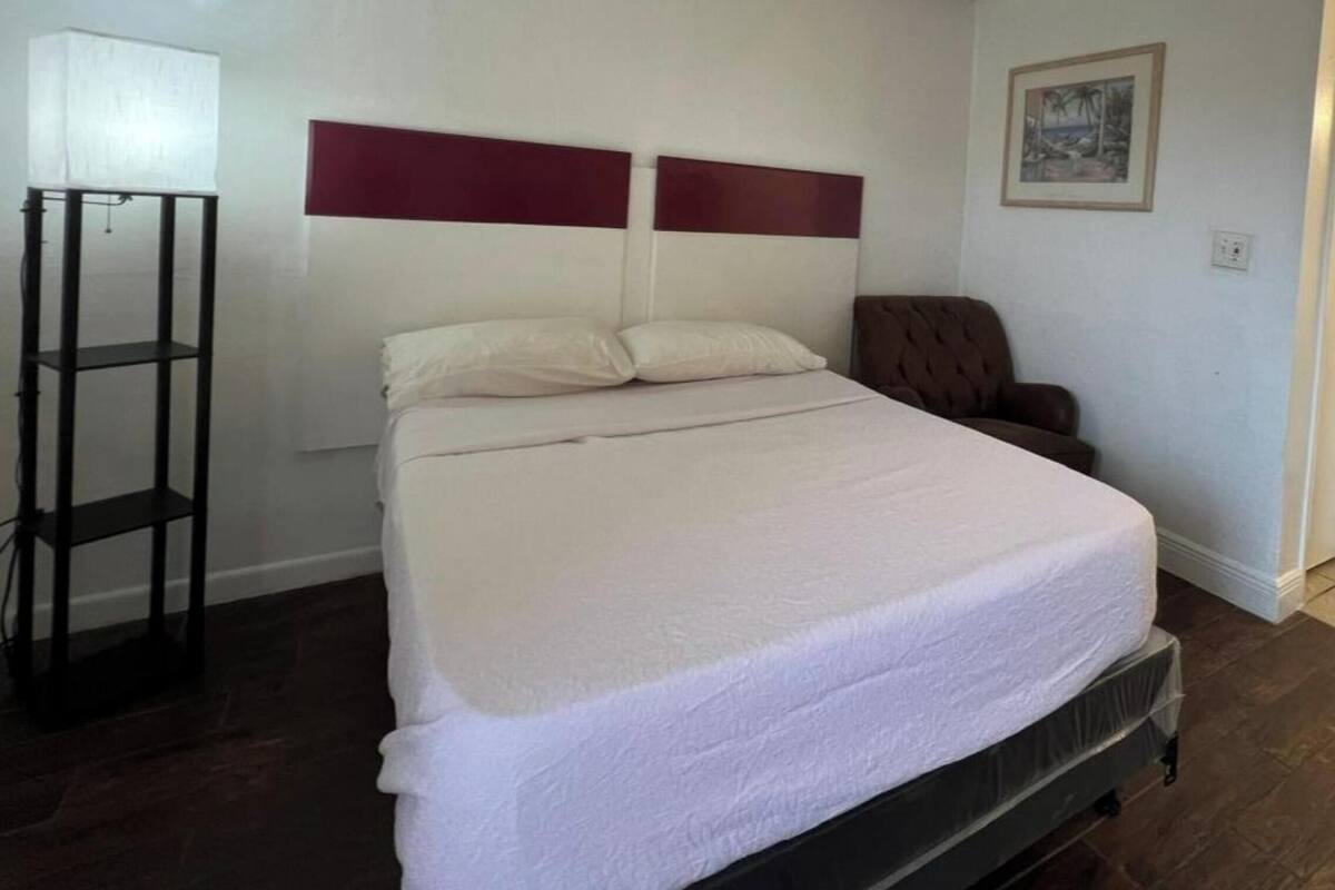A single bed with a white mattress and headboard, a bedside table, a chair, and a standing lamp.