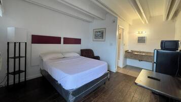 A bedroom with a bed, a desk, a chair, a TV, and a bathroom with a mirror and sink.