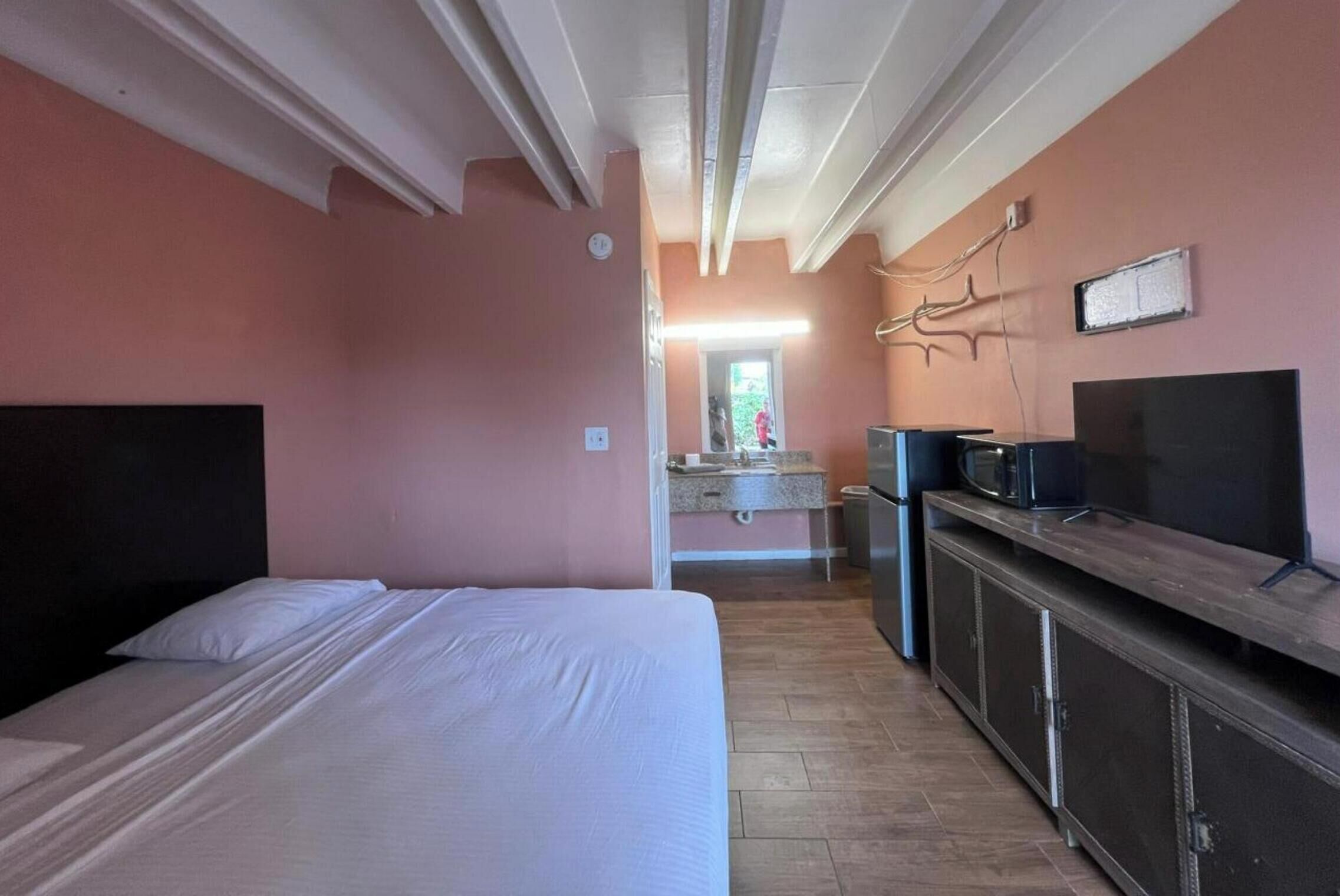 A hotel room with a bed, a TV, a kitchenette, and a bathroom visible in the background.