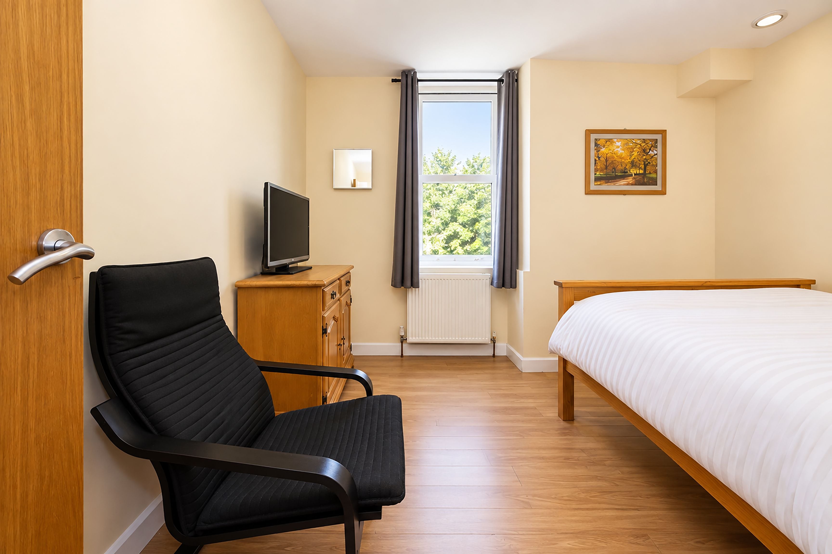 A hotel room with a bed, a chair, a television, a window with curtains, and a painting on the wall.