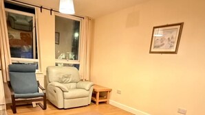 Een woonkamer met een houten vloer, een beige bank, een blauwe fauteuil, een klein houten tafeltje en een vloerkleed met geometrische patronen.