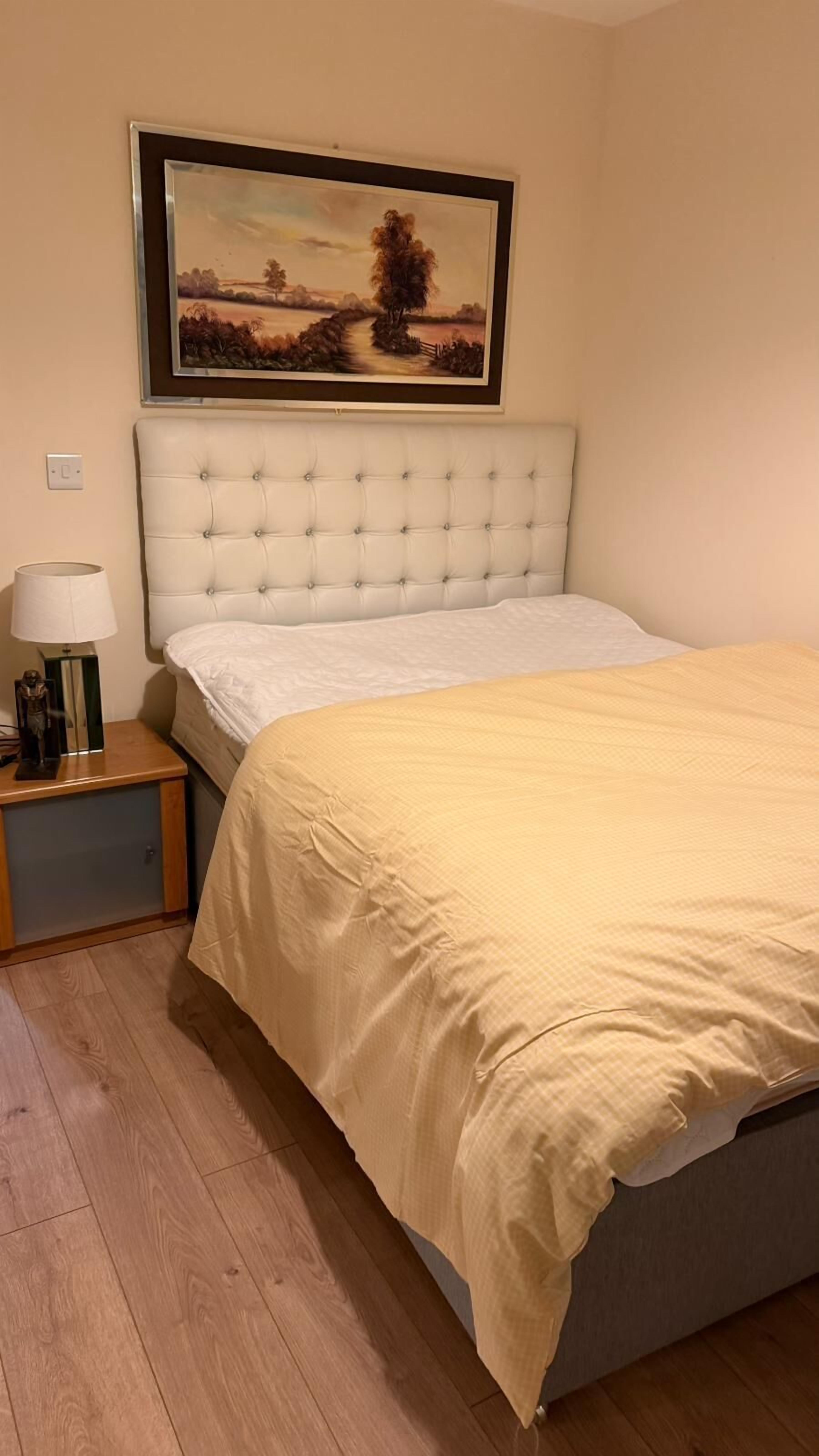 A neatly made bed with a beige bedspread, a white headboard, and a bedside table with a lamp and a small statue.