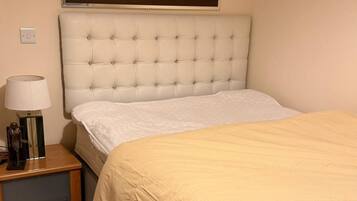 A neatly made bed with a beige bedspread, a white headboard, and a bedside table with a lamp and a small statue.
