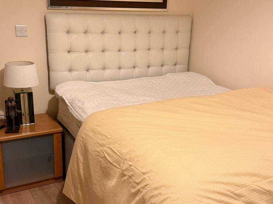 A neatly made bed with a beige bedspread, a white headboard, and a bedside table with a lamp and a small statue.