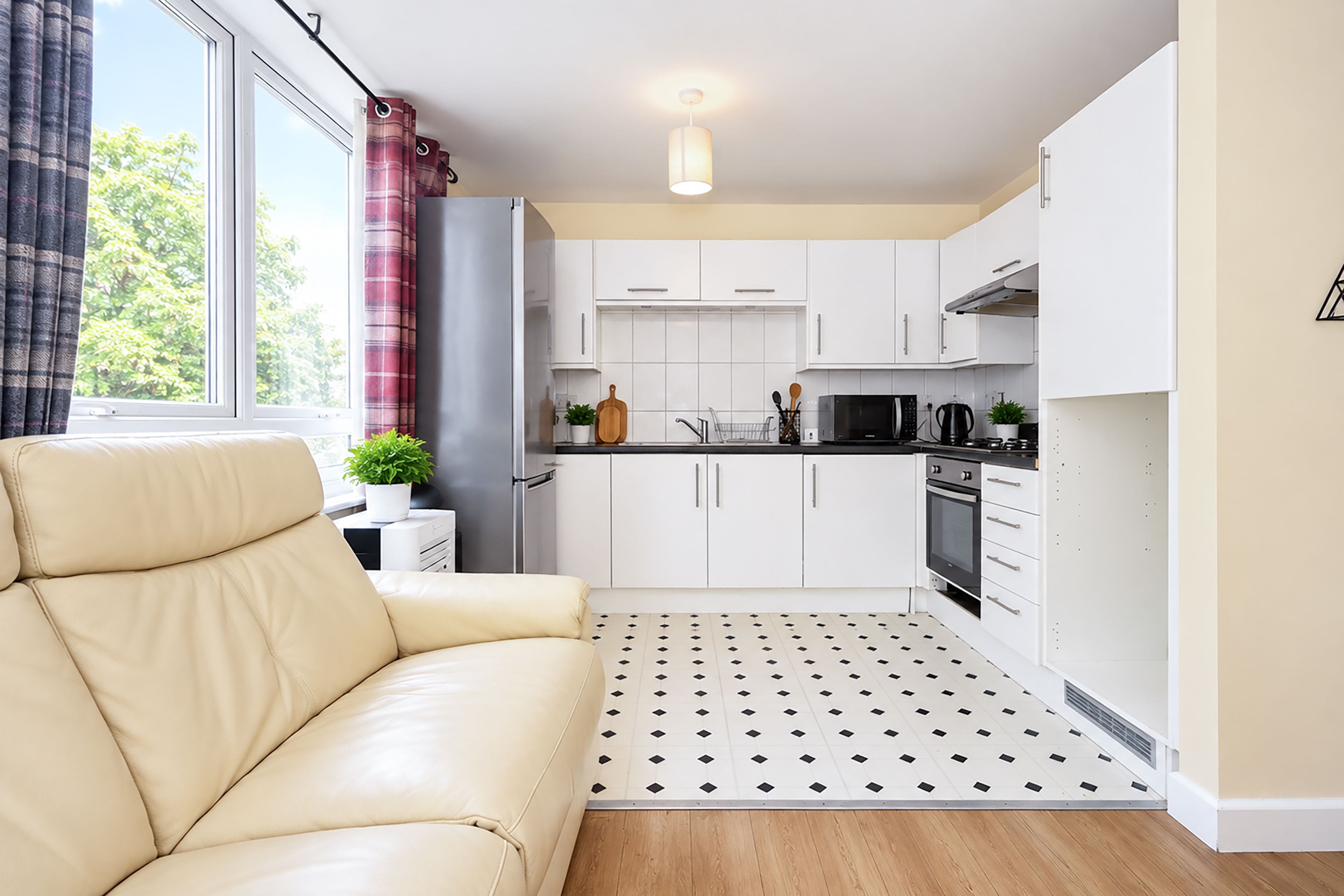 Une cuisine compacte avec des meubles blancs, un plan de travail noir et un four à micro-ondes. On y trouve un canapé beige et une fenêtre avec des rideaux.