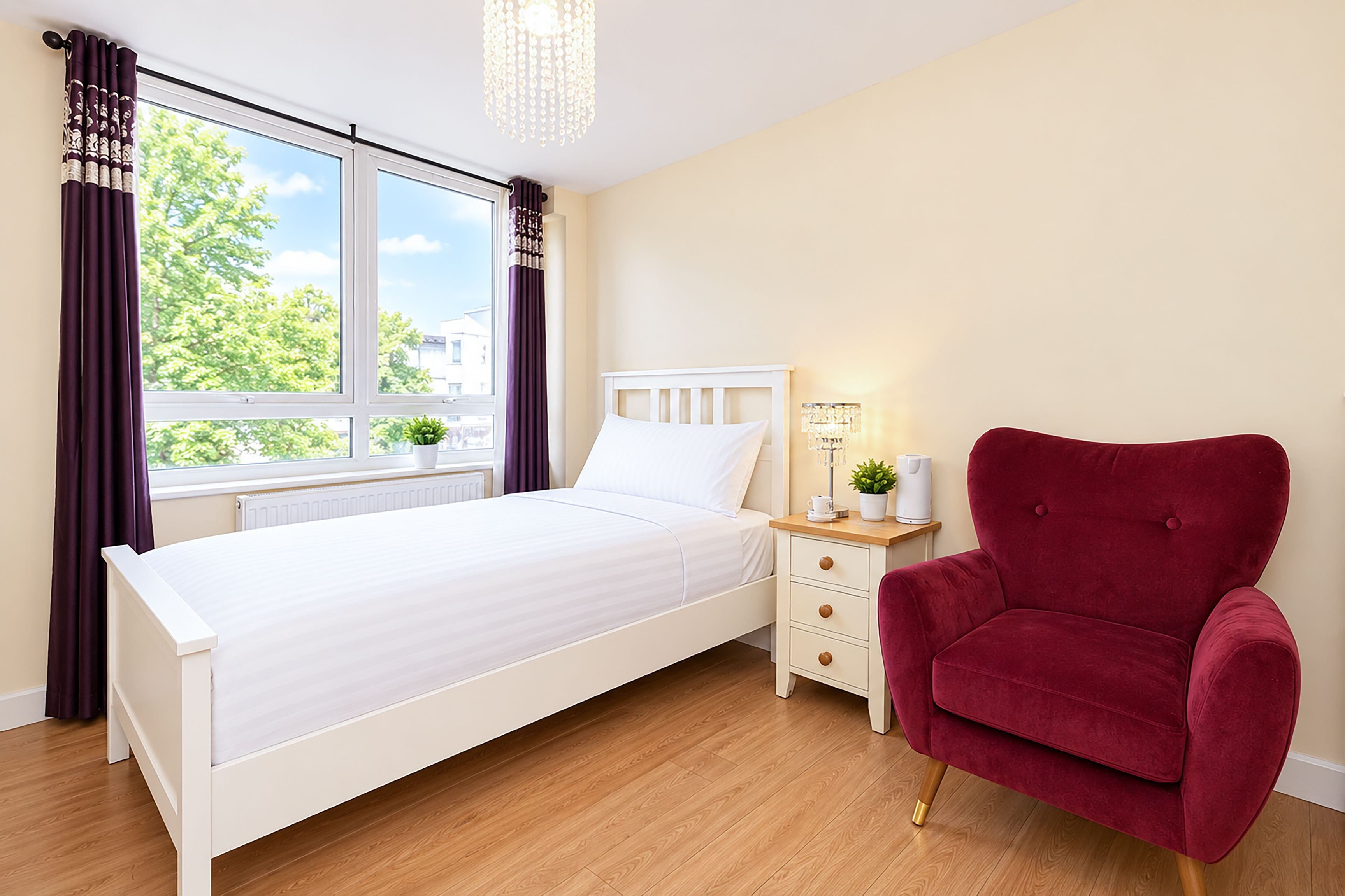 A bedroom with a bed, a nightstand, a red chair, and a window with curtains.