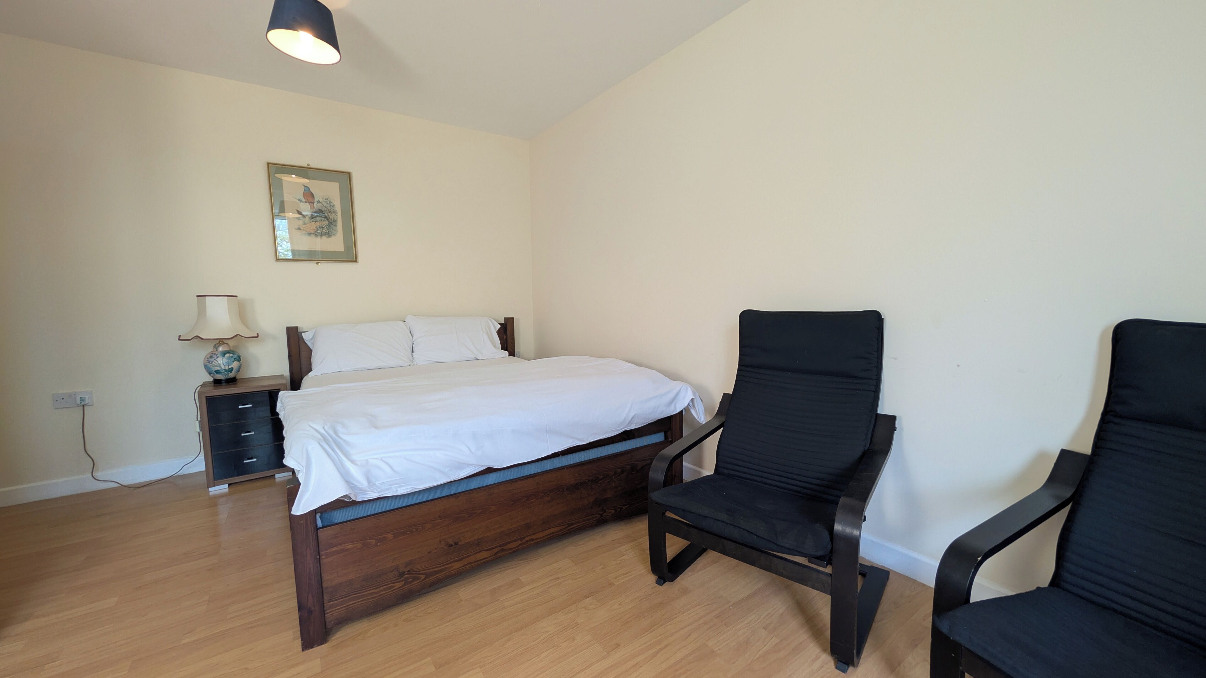 Un dormitorio con cama de madera, una mesita de noche, una lámpara, una silla y un cuadro colgado en la pared.