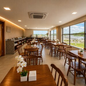 A spacious dining area with multiple tables and chairs, a kitchen area with buffet setup, and large windows offering a view of the outside.