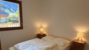 Una cama individual con cabecera de madera, dos mesitas de noche con lámparas y una ventana con vistas a un paisaje montañoso.