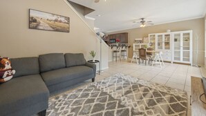 Ein Wohnzimmer mit Sofa, Teppich, Gemälde, Deckenventilator, Treppe, Küchenbereich und Esstisch.