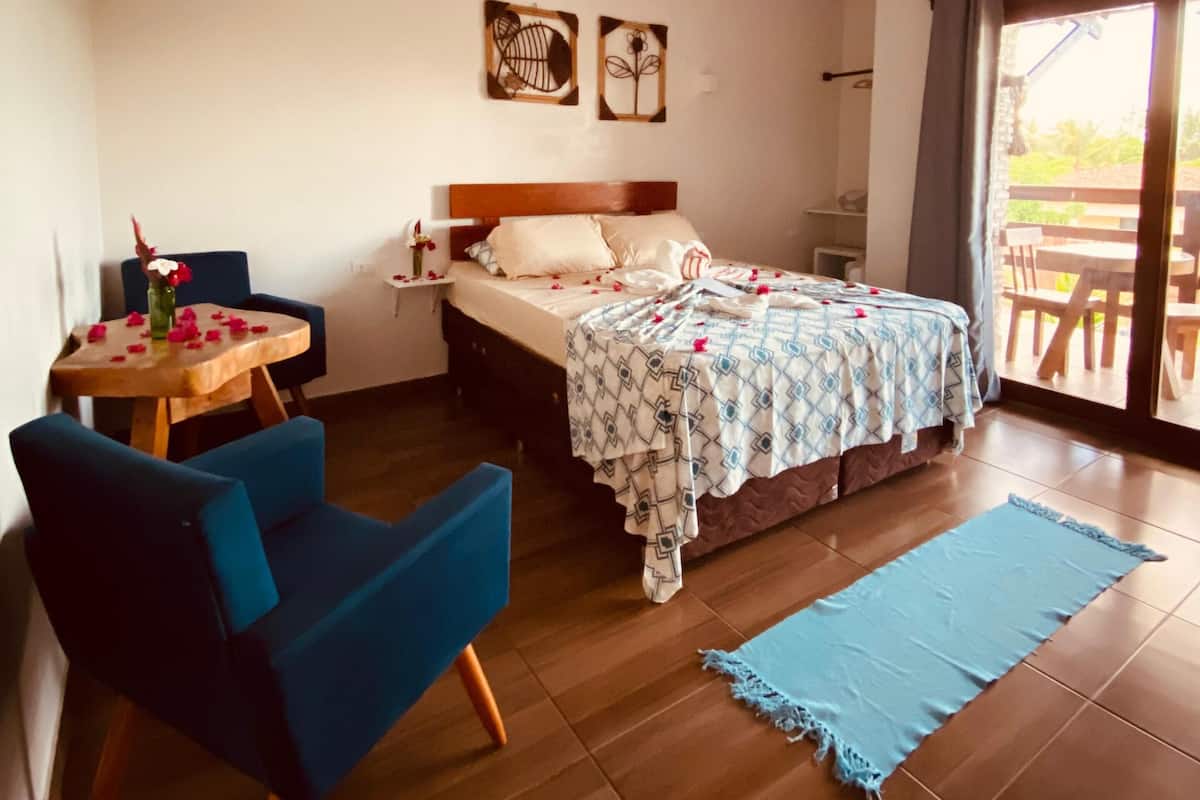 A bedroom with a bed, a table with chairs, a blue chair, a wooden floor, and a view through a glass door.