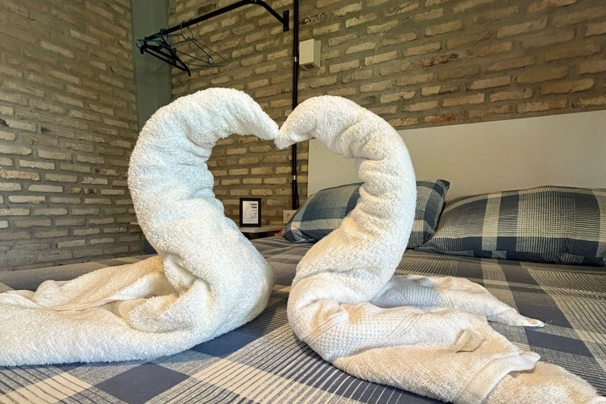 A hotel room with a brick wall, a bed with a checkered blanket, and two towels arranged in a heart shape.