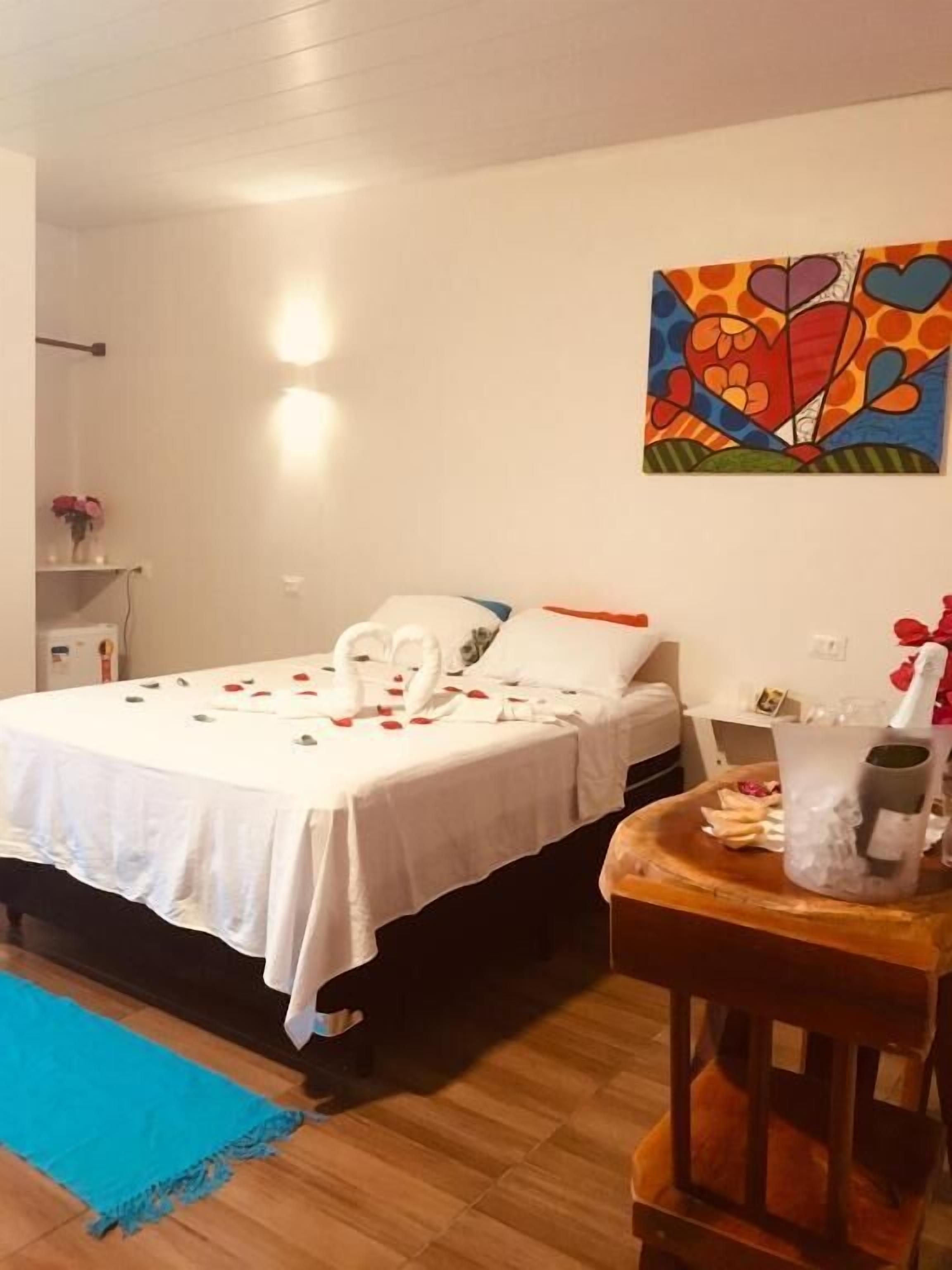 A neatly made bed with a white bedspread, a bouquet of roses, and a heart-shaped decoration on the headboard. A colorful abstract painting adorns the wall above the bed.