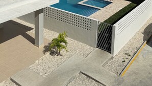 Et moderne poolområde med en rektangulær pool, en lille hvid bygning med et skilt, og udsigt over et grønt landskab med træer og bygninger i baggrunden.