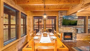 Ein Esszimmer mit Holzboden, einem Kamin aus Stein, großen Fenstern und einem an der Wand befestigten Fernseher.