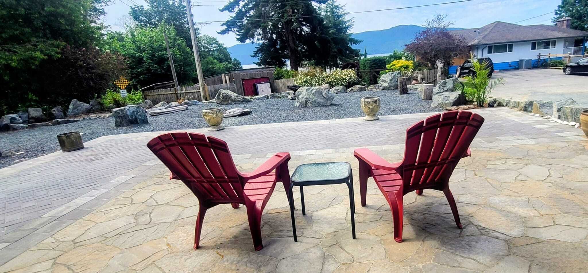 Two red Adirondack chairs and a small table on a stone patio with a view of a mountain and a house in the background.