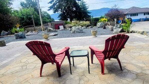Two red Adirondack chairs and a small table on a stone patio with a view of a mountain and a house in the background. - MALASPINA HIDEAWAY 2 (Powell River)