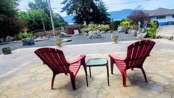 Two red Adirondack chairs and a small table on a stone patio with a view of a mountain and a house in the background.