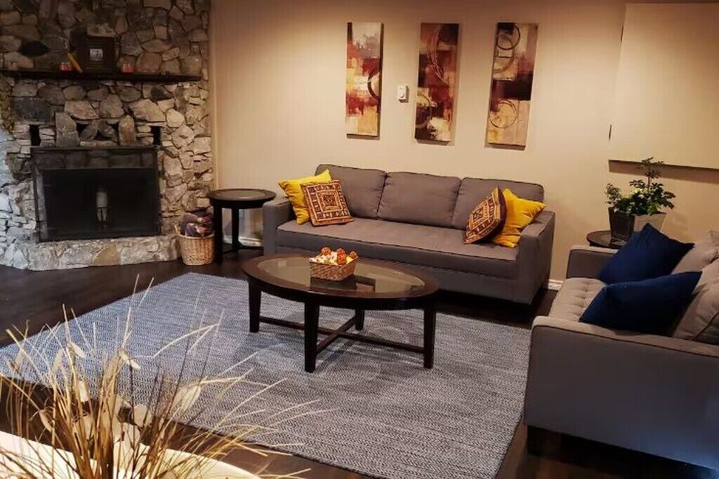 A cozy living room with a stone fireplace, a grey sofa with yellow cushions, a round coffee table, and a grey armchair.