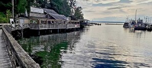 Un restaurante frente al agua con un muelle que se adentra en aguas tranquilas, rodeado de barcos y árboles.