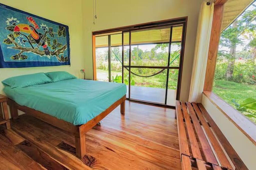 Une chambre à coucher avec un lit, un banc en bois, une grande fenêtre et un tableau coloré au mur.