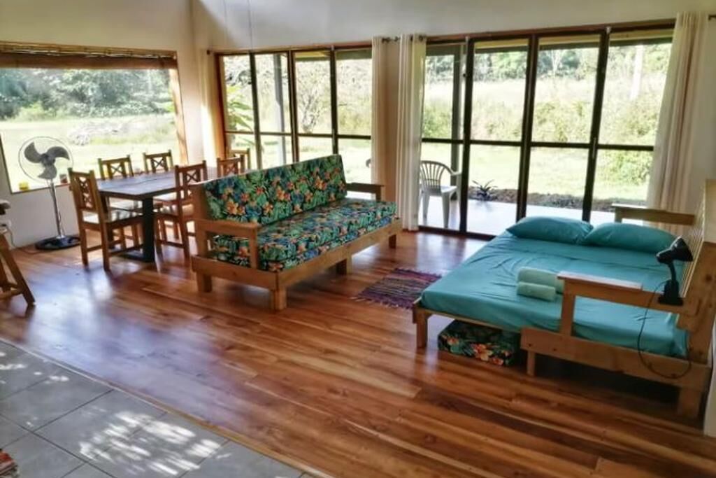 Une pièce dotée d’un plancher en bois, d’un canapé à motifs floraux, d’un lit recouvert d’une courtepointe turquoise et de grandes fenêtres donnant sur un paysage verdoyant.