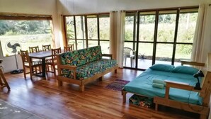 Ein Zimmer mit Holzboden, einer Couch mit Blumenmuster, einem Bett mit türkisblau versteckter Bettdecke und großen Fenstern mit Blick auf Grünflächen.