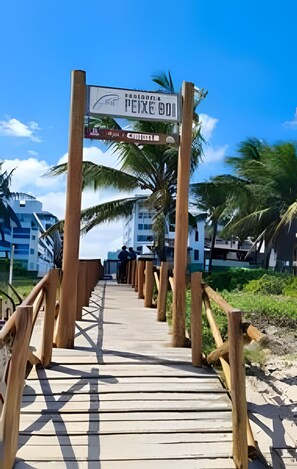 A wooden boardwalk leading to a hotel entrance with palm trees and modern buildings in the background. - 1503 Apto 2 Qtos completo João Pessoa (Cabedelo)