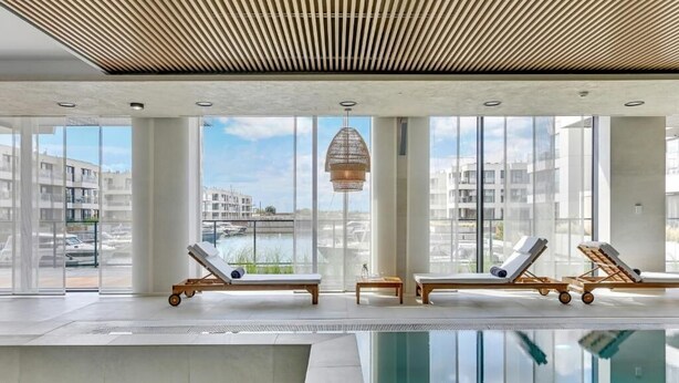 Un área de piscina interior moderna con paredes de vidrio transparente, sillas de descanso y una lámpara en el techo.