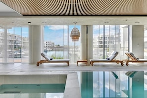 Un área de piscina interior moderna con paredes de vidrio transparente, sillas de descanso y una lámpara en el techo.