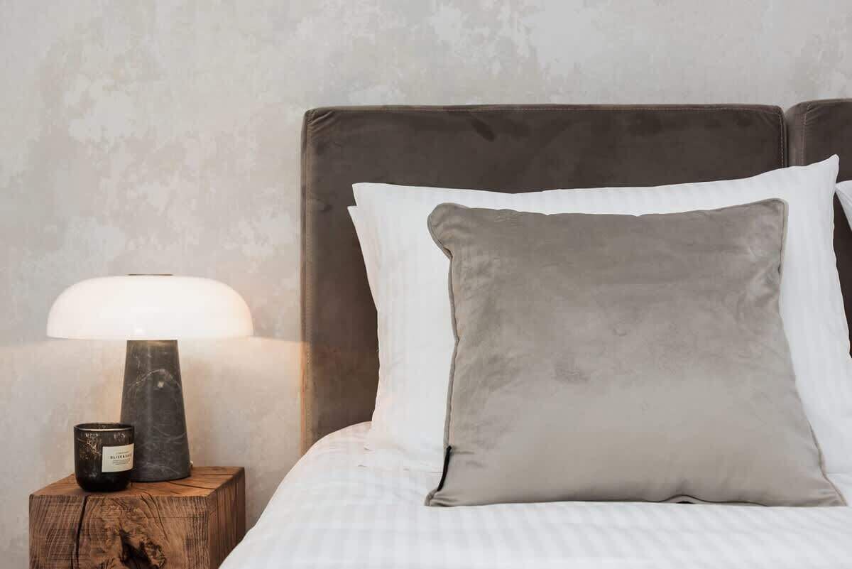 A neatly made bed with a grey headboard and white pillows, a bedside table with a lamp and a candle, and a textured wall in the background.