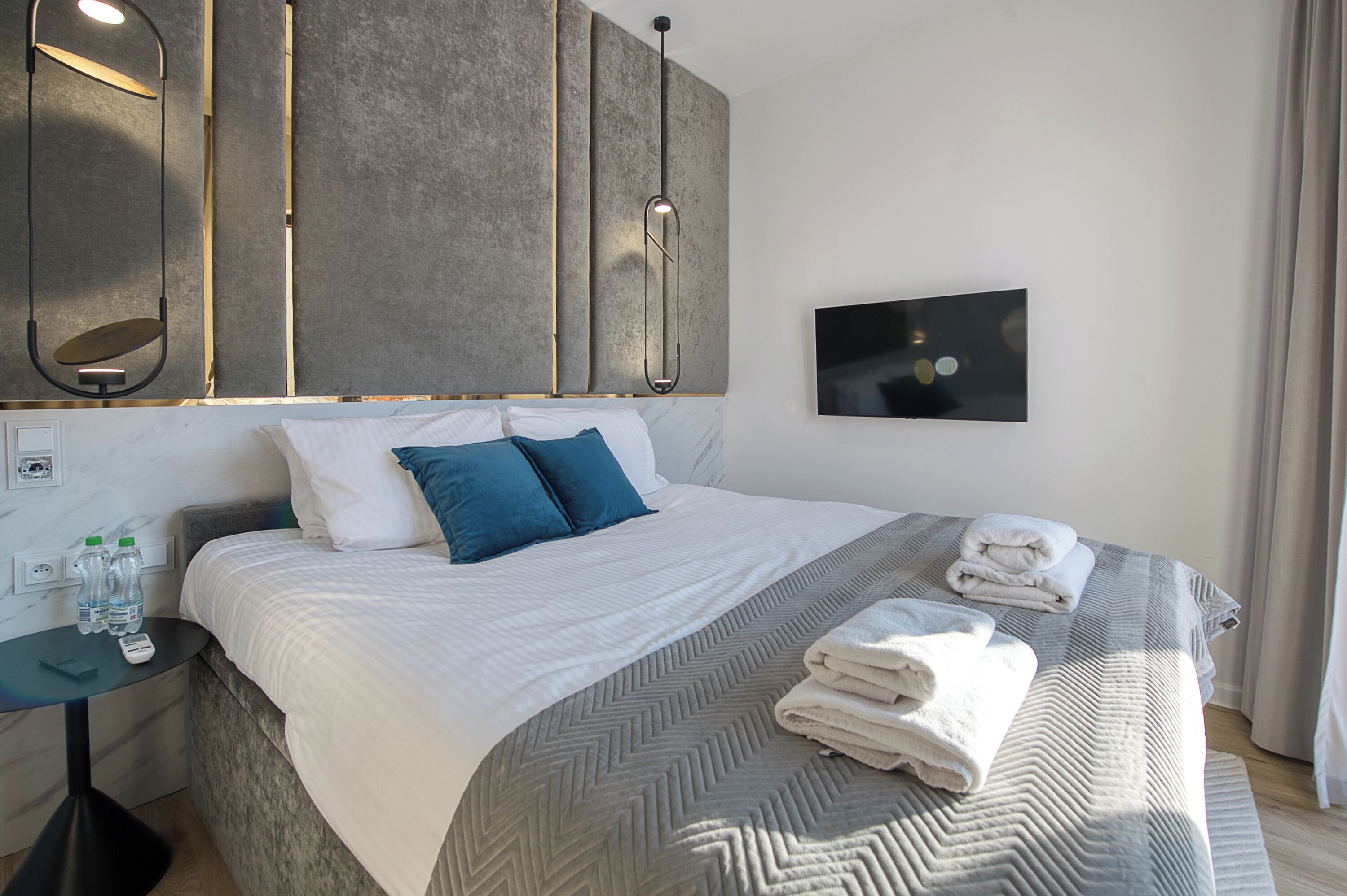 Una habitación de hotel moderna con una cama grande, una mesita de noche con botellas de agua, un televisor de pantalla plana y un espejo amplio.