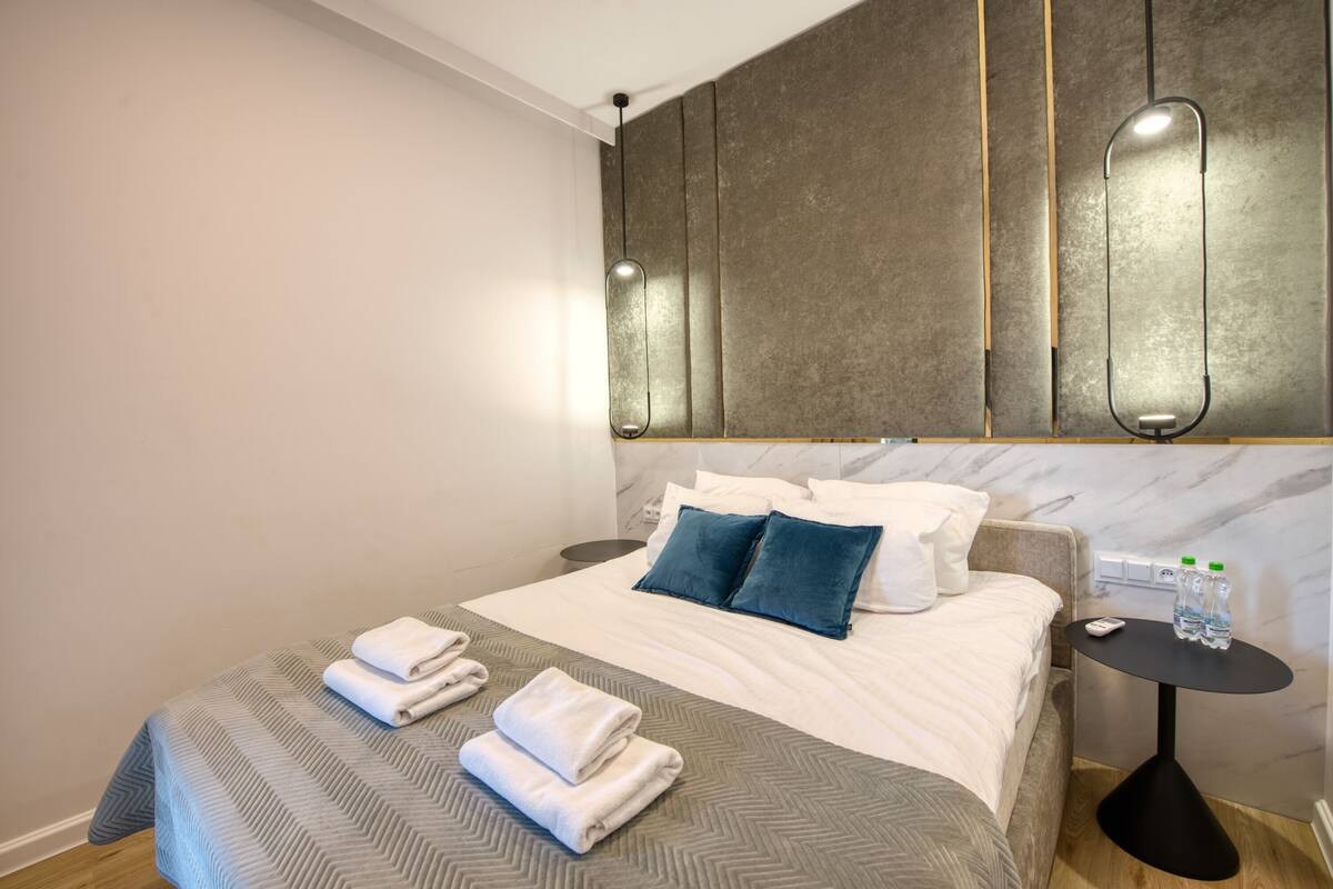 A modern hotel room with a large bed, two pillows, a bedside table with water bottles, and a wall-mounted light fixture.