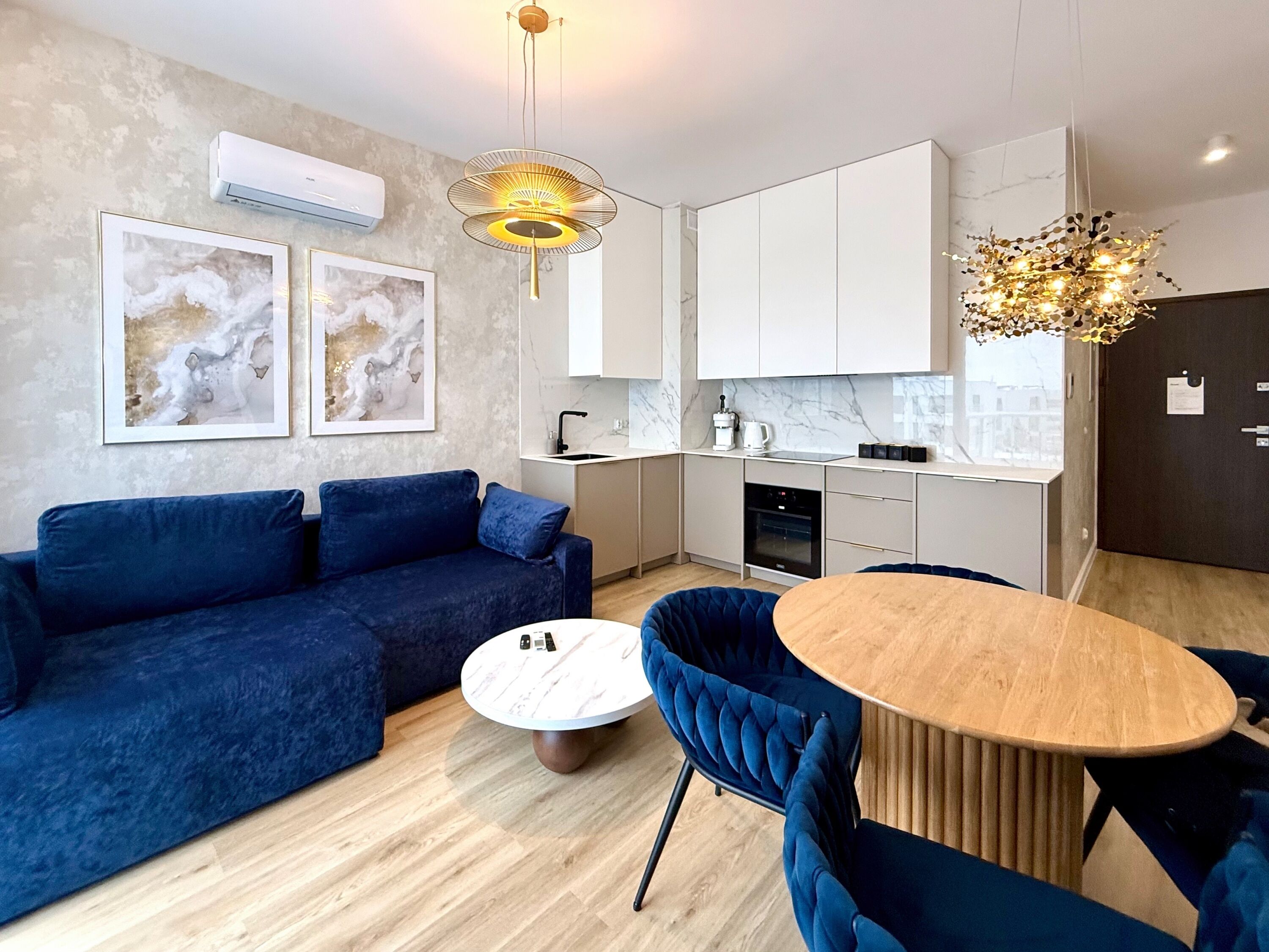 A modern living room with a blue sofa, a round wooden coffee table, and a dining area with a wooden table and blue chairs. There is a kitchen with white cabinets and a marble backsplash.