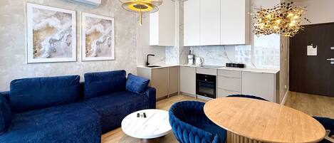 A modern living room with a blue sofa, a round wooden coffee table, and a dining area with a wooden table and blue chairs. There is a kitchen with white cabinets and a marble backsplash.