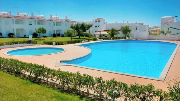 A swimming pool with a circular design, surrounded by a paved deck. There are white buildings with red roofs in the background. - Quinta do Torrejão 800m plaga Armação de pêra (Porches)