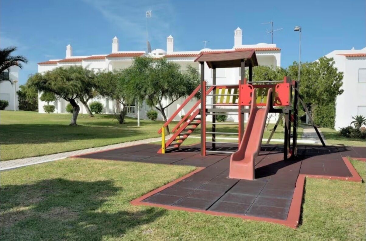 Ein Spielplatz mit einer roten Rutsche und Treppen, umgeben von Gras und Bäumen, mit Gebäuden im Hintergrund.