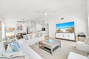 Un salon moderne avec un canapé blanc, une table basse et un grand téléviseur diffusant une scène de plage.