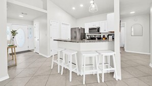 A modern kitchen with white cabinetry, a stainless steel refrigerator, a microwave, and a granite countertop. There are four white bar stools and a hanging pendant light. - Destin/Miramar Beach Escape, Family-Friendly + Private Beach Access (Destin)