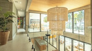 A modern hotel lobby with a large chandelier, glass railings, a central island, and a view of palm trees through large windows.