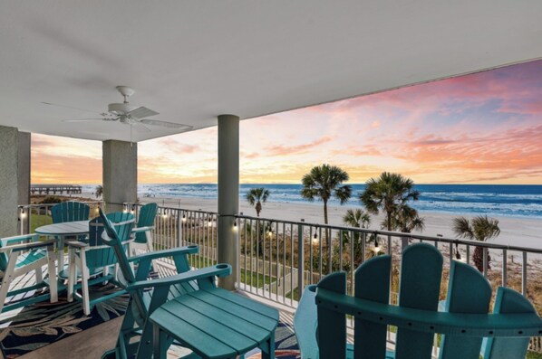 A balcony with teal chairs and a table, overlooking a beach with palm trees and a sunset. - Beachside Haven - enjoy 3 beachfront bedrooms, 2nd floor, free beach chairs (Panama City Beach)