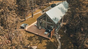 Une cabane moderne dotée d’une terrasse en bois, de grandes fenêtres et d’un toit en métal, entourée d’arbres et d’un paysage rocheux.