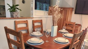 A dining area with a wooden table set for six, surrounded by wooden chairs with black cushions. A white vase with dried flowers is on the table, and a potted plant is on the counter. - Orange Grove Homestead - Spacious Garden Holiday Home (Kerikeri)