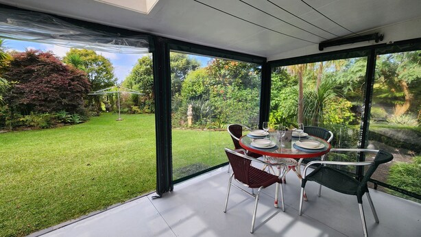 A conservatory with a glass ceiling, a round table with chairs, and a view of a well-maintained garden. - Orange Grove Homestead - Spacious Garden Holiday Home (Kerikeri)