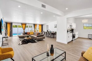 A modern living room with a sofa, coffee table, and dining area. There is a kitchen with white cabinets and a dining table. - LUXURY POOL VILLA 5BD 4BR #3, Heated Pool (Miami)