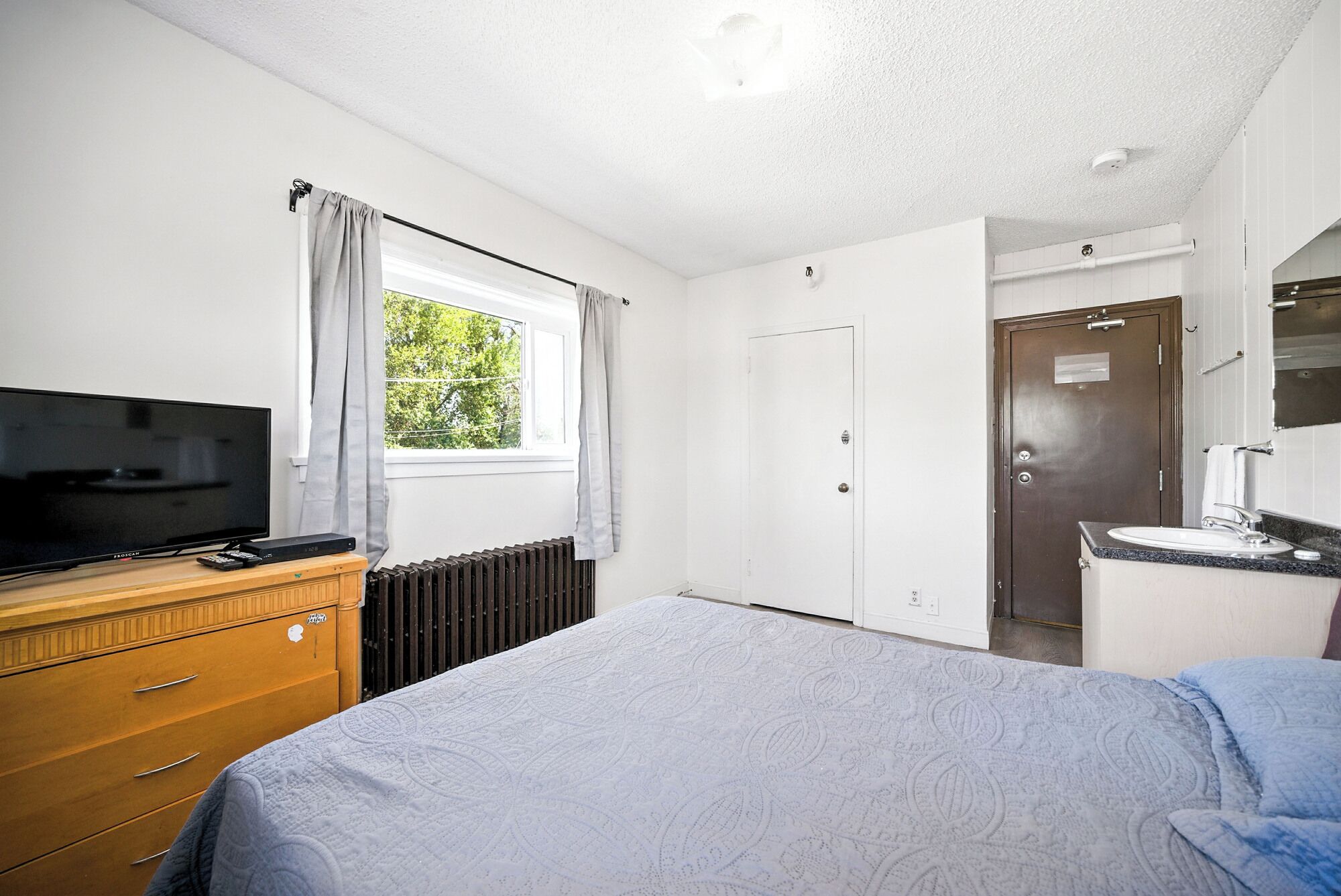 Un dormitorio con cama, cómoda, televisor, ventana con cortinas, radiador, puerta y baño con lavamanos y espejo.