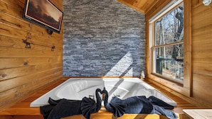 A wooden cabin with a stone wall, a large window, and a bathtub with towels. - Smoky Mountain Views • Hot Tub • Fireplace • Cozy Cabin (Sevierville)