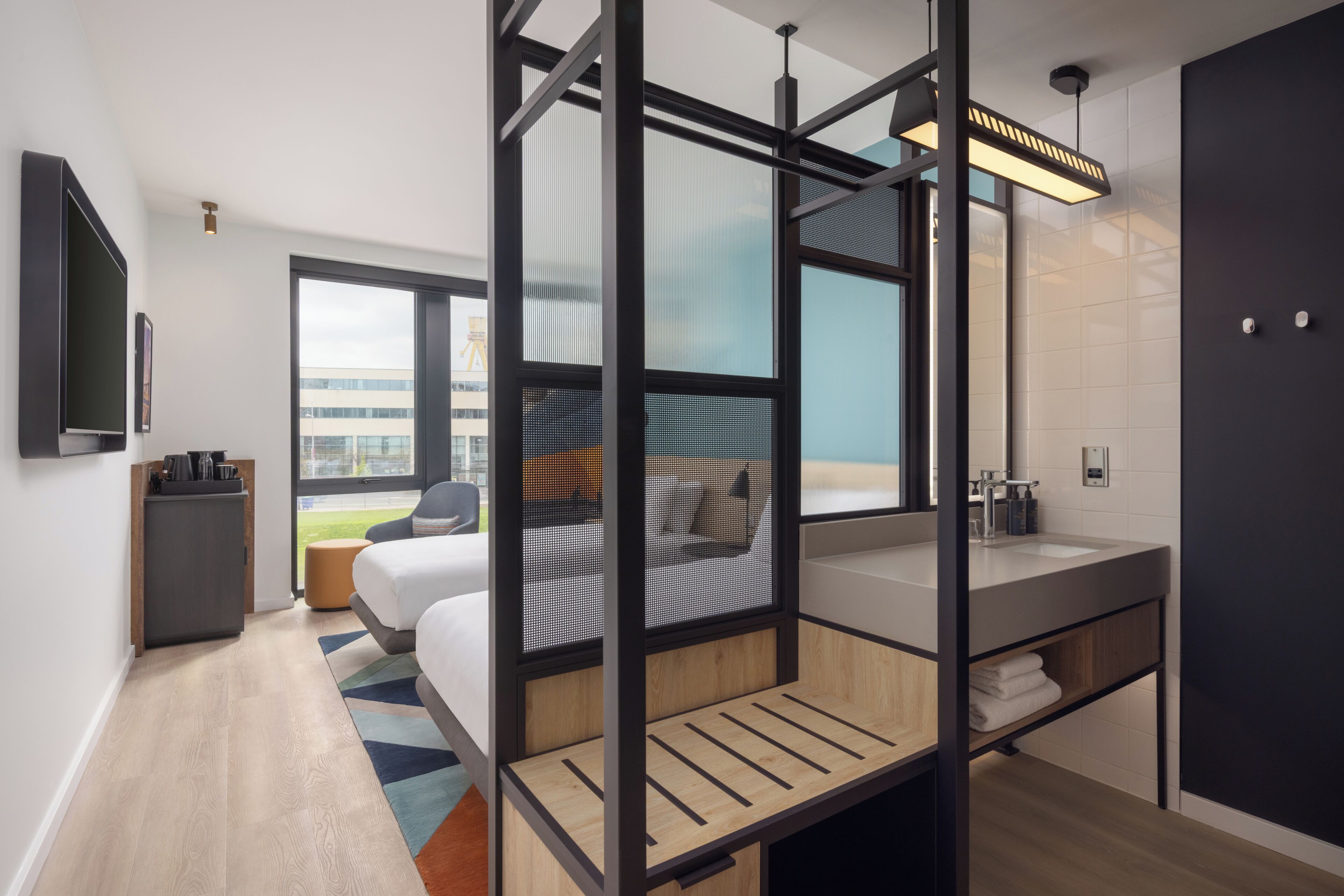 Quarto de hotel moderno com cama, escrivaninha, janela ampla, banheiro com pia e vista para o exterior.