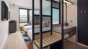 A modern hotel room with a bed, a desk, a large window, a bathroom with a sink, and a view of the outside.